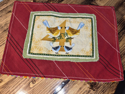 Red fabric with a embroidered design of 4 calling birds and tree on a wooden surface